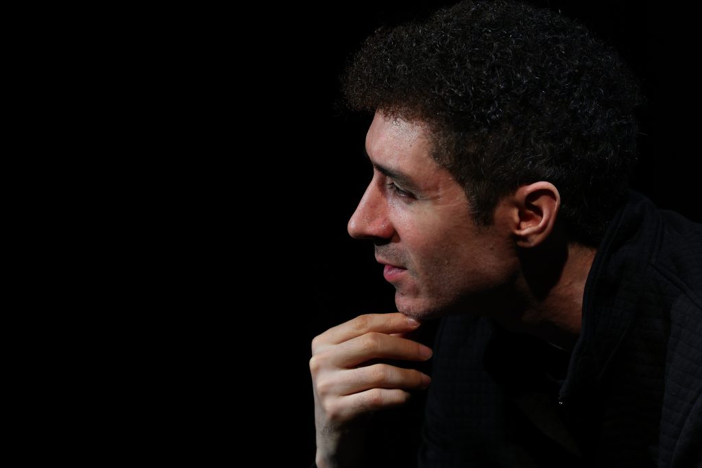 Side profile of a man with curly hair in deep thought, resting his chin on his hand. The image is dramatically lit, with the subject illuminated against a dark background, emphasizing the thoughtful expression on his face.