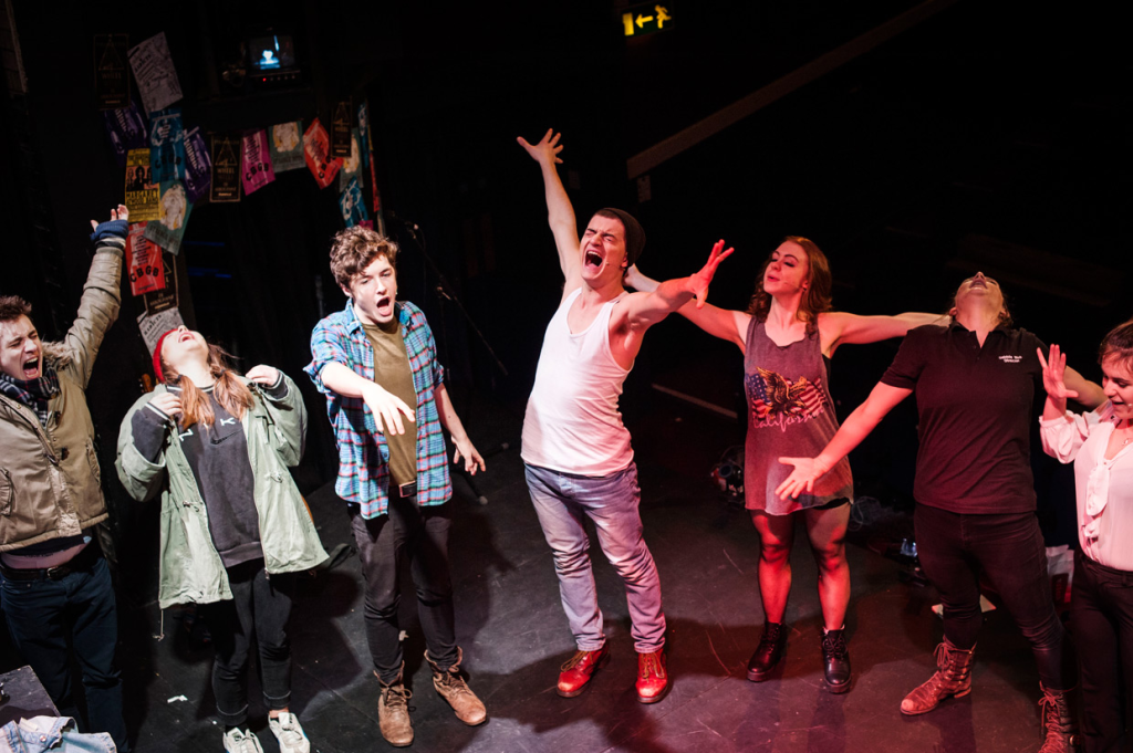 This image shows a group of seven people, likely actors, standing on a stage in a theatrical environment. They are in the middle of an exaggerated, expressive movement, with their arms stretched out and faces animated, possibly during a warm-up or rehearsal. The individuals are dressed casually, with a variety of clothing styles, including a plaid shirt, tank top, and boots. The background includes posters and flyers, adding to the informal, energetic, and creative atmosphere of the space. The scene reflects the intensity and excitement typically seen in theater practice or an ensemble performance moment.