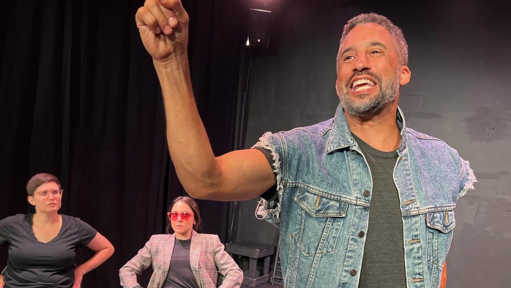 This image shows a stage scene where three actors are performing. In the foreground, a man with short, graying hair and a beard is energetically pointing upward with a confident expression. He is wearing a sleeveless, frayed denim jacket over a dark t-shirt. Behind him, seated, are two other performers. One is dressed in casual jeans, a black t-shirt, and glasses, with their arms resting on their hips. The other is wearing sunglasses, a plaid blazer, jeans, and sneakers, sitting with legs crossed in a more relaxed pose. The stage lighting is bright, illuminating the actors against a dark backdrop.