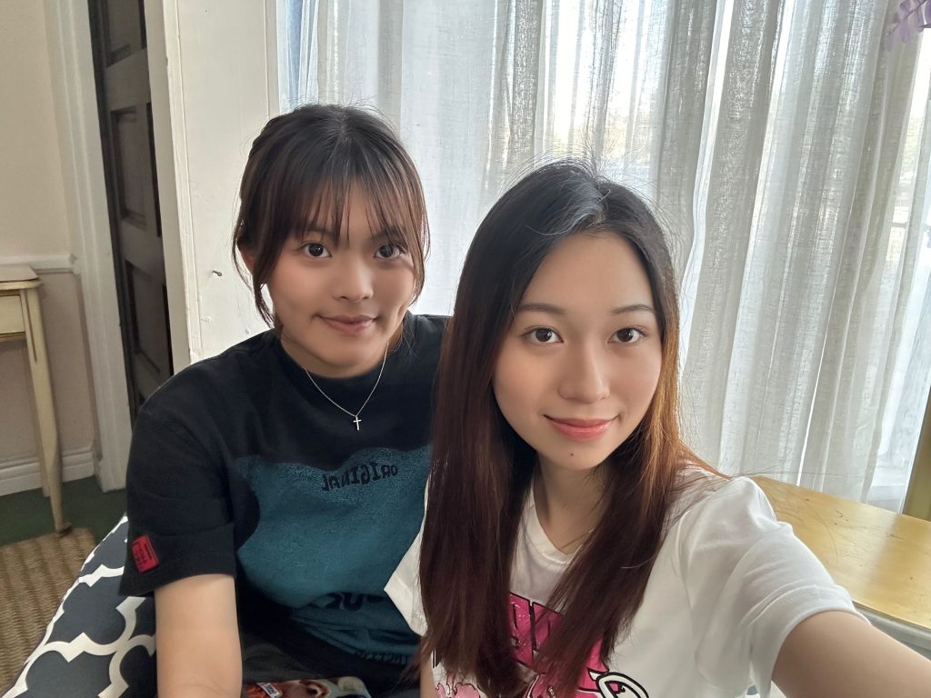 Two people sitting indoors, smiling at the camera. One wears a dark shirt, and the other a white shirt. Light curtains are in the background.