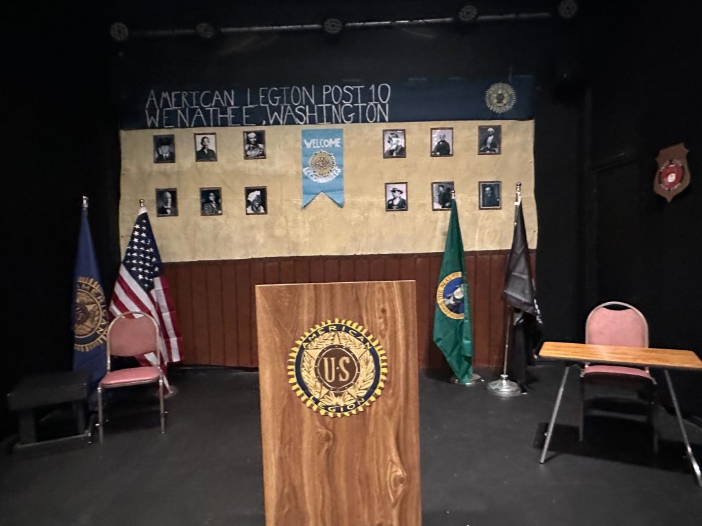 This image appears to depict a set design for a stage production. The backdrop is styled as a veterans' hall with the banner reading "American Legion Post 10, Wenatchee, Washington." Several framed photographs of military figures are arranged on the wall, and there are U.S. and Washington state flags on display. A wooden podium with the U.S. Legion emblem stands prominently in the foreground, with two empty chairs placed on either side, creating a formal, institutional atmosphere. The setup suggests that this scene could be part of a performance involving themes related to veterans, American history, or constitutional rights.