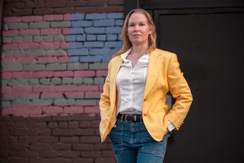 The image depicts a confident woman standing outdoors in front of a brick wall painted with a faded, stylized American flag. She is wearing a yellow blazer over a white button-up shirt and blue jeans, with her hands in her pockets. The woman is looking directly at the camera, exuding a composed and strong demeanor. The backdrop suggests an urban setting with muted tones contrasting the bright yellow jacket.