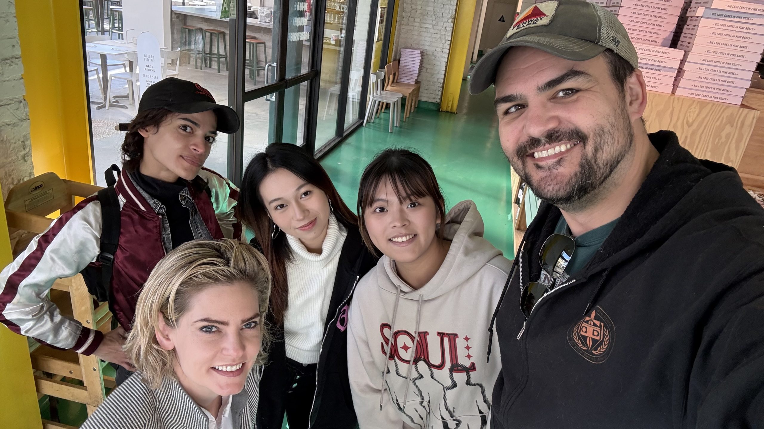 Group photo of five people smiling indoors. The group includes four women and one man, all casually dressed. The setting appears to be a brightly lit cafe or restaurant with green flooring and wooden furniture in the background.