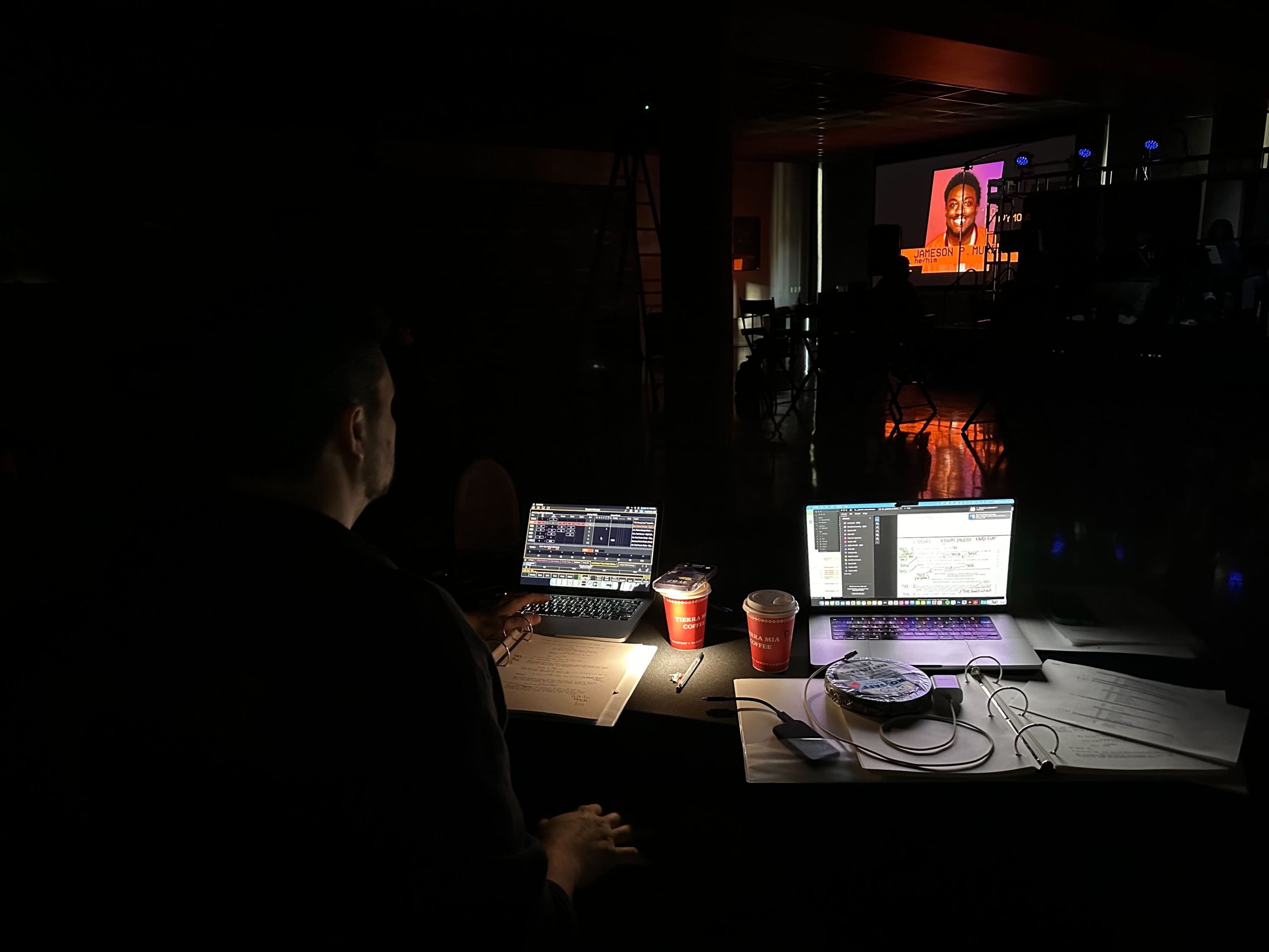 "A dimly lit workspace with a person seated at a desk operating two laptops. The desk is cluttered with papers, cables, a wrapped dish, and two coffee cups. In the background, a large screen displays a presentation featuring a person's photo and name, with faint silhouettes of other people and equipment visible."