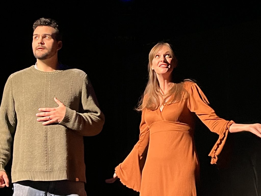 Two actors stand on stage in dramatic lighting. The man on the left, wearing a green knit sweater, looks thoughtful with his hand resting on his chest. The woman on the right, dressed in a burnt orange dress with flowing sleeves, gazes upward with an expression of wonder or hope. Both are bathed in warm light, standing against a dark background.