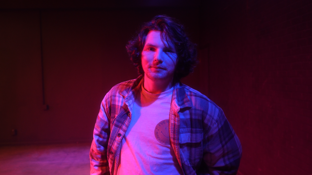 A young man with wavy, shoulder-length hair stands in a dimly lit room with a moody red and blue lighting effect. He wears a plaid shirt over a light-colored T-shirt with a faint circular design. His expression is neutral, and he looks directly at the camera. The background is dark with a hint of an industrial or warehouse-like setting.