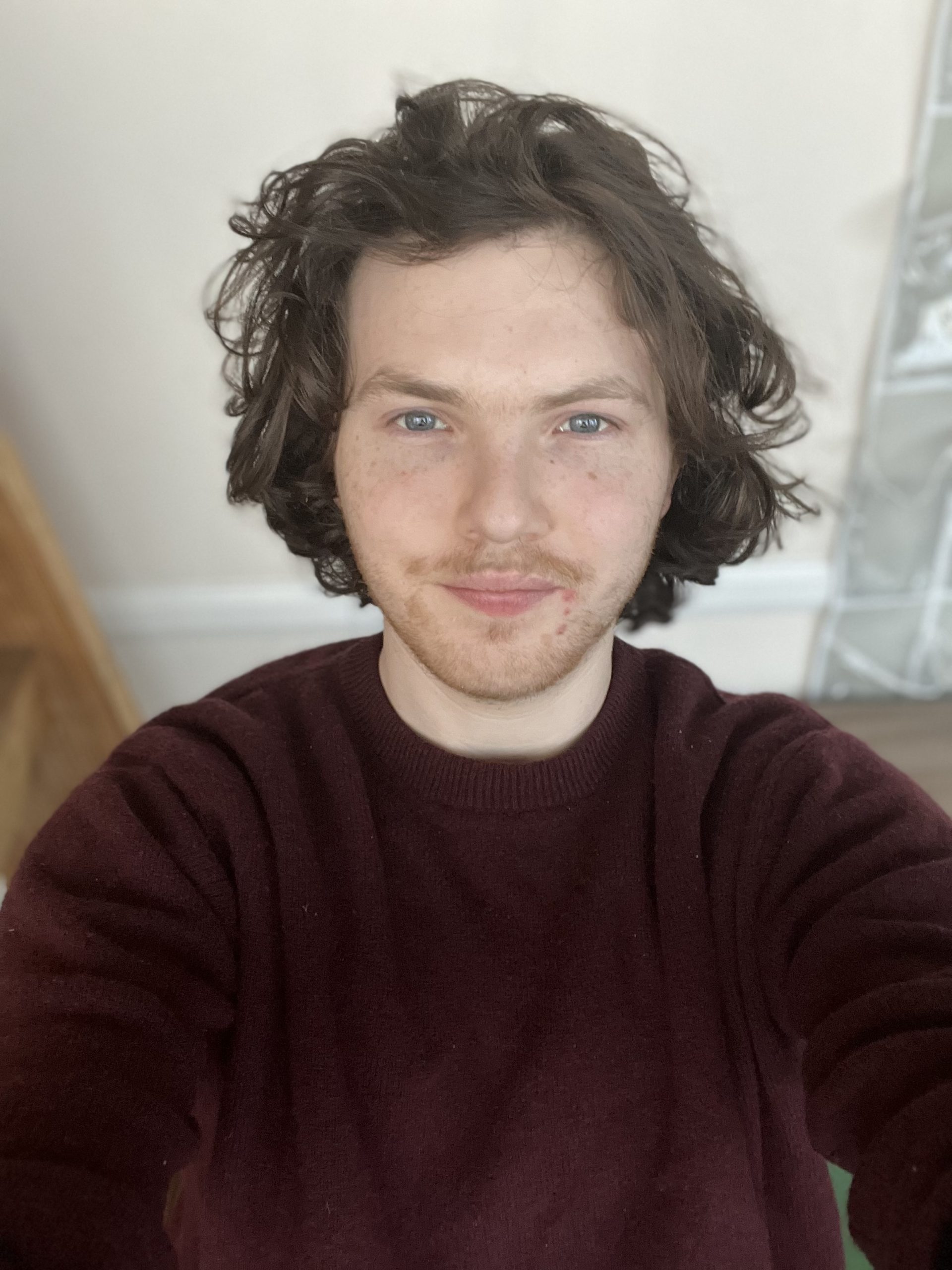 A young man with wavy, shoulder-length brown hair and blue eyes takes a selfie indoors. He has a light beard and mustache and is wearing a burgundy sweater. His expression is relaxed with a slight smile. The background features a wooden staircase, a white wall, and part of a reflective surface, possibly a window or mirror. The lighting is soft and natural.