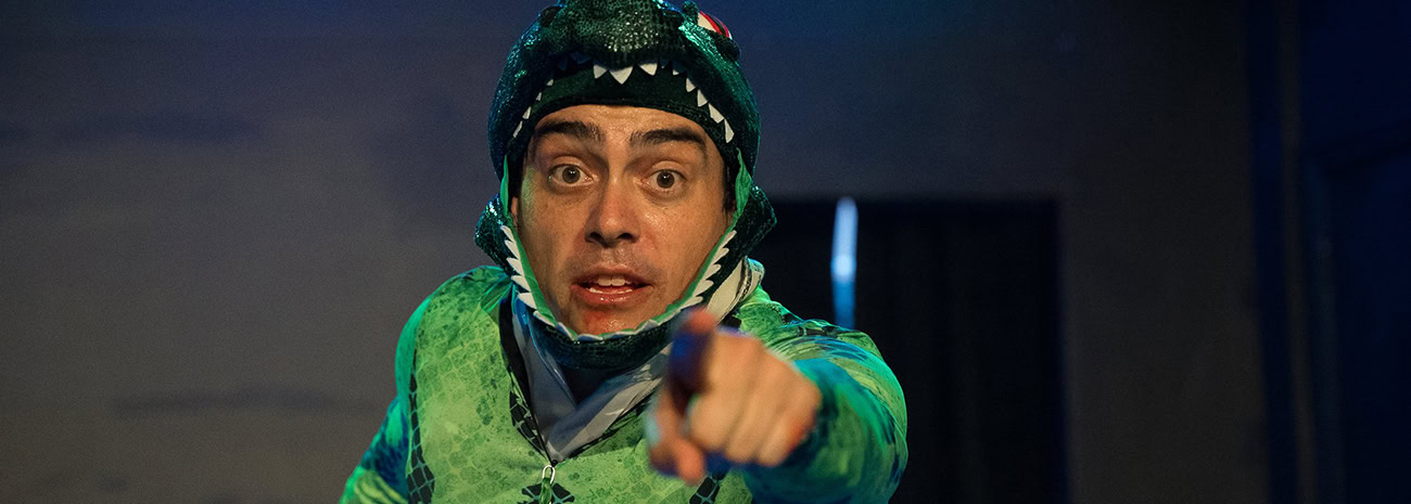 A man dressed in a green dinosaur costume with a hood featuring teeth and eyes is captured in an intense moment. His facial expression is animated, with wide eyes and an open mouth, and he is pointing directly at the camera, creating a sense of urgency or engagement. The dimly lit background suggests a theatrical or performance setting, enhancing the dramatic atmosphere of the image.