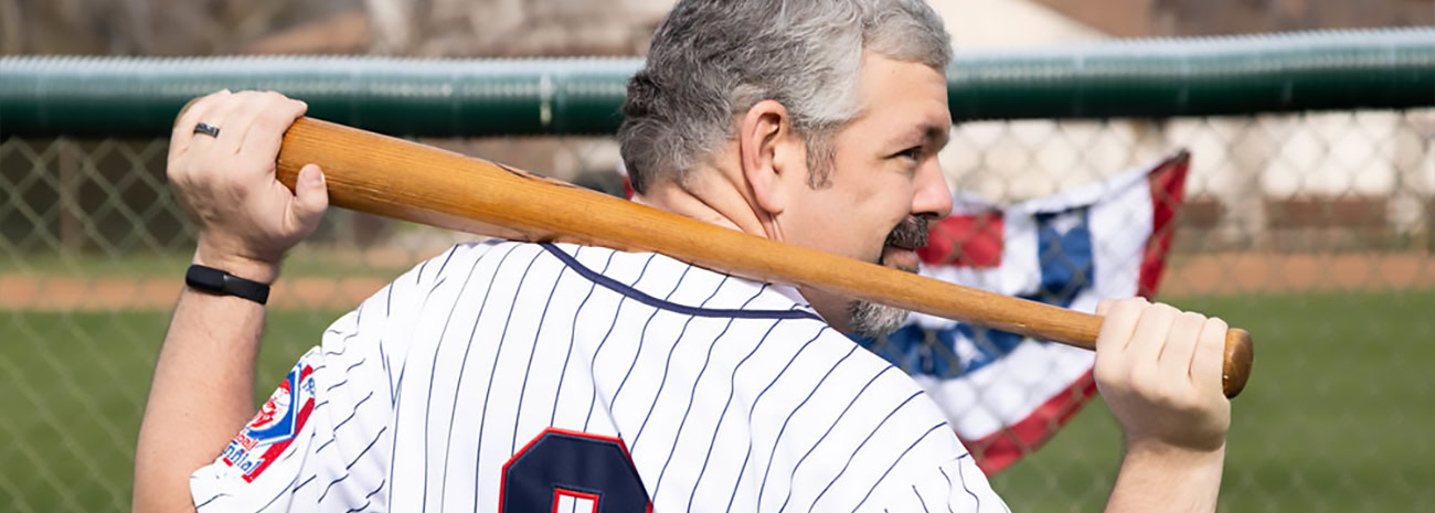 A man in a vintage-style baseball jersey, featuring blue pinstripes and the number "9," stands in a baseball field holding a wooden bat over his shoulder. He faces slightly to the side, showing a focused expression as he prepares for the game. In the background, a baseball field fence is visible, with a partially visible red, white, and blue flag fluttering in the breeze. The image captures a nostalgic and energetic baseball vibe.