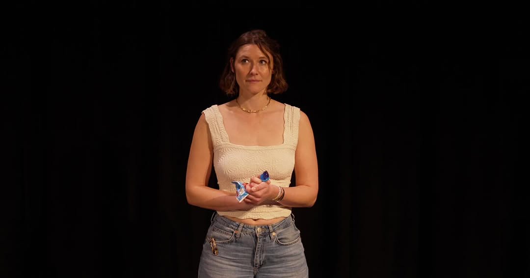 A woman with short brown hair and light skin stands on a dimly lit stage against a black background. She is wearing a cream-colored, sleeveless top and blue jeans.