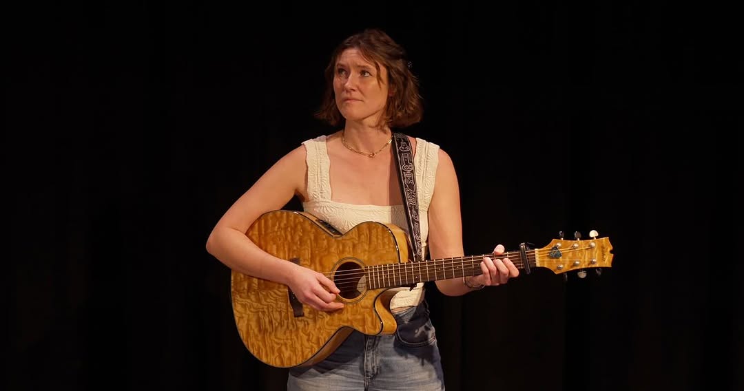 A woman with short brown hair and light skin stands on a dimly lit stage against a black background, holding an acoustic guitar. She is wearing a cream-colored, sleeveless top and blue jeans.